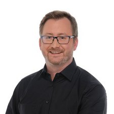 Mann mit Brille und freundlichem Lächeln vor hellem Hintergrund In einem hellen Raum steht ein Mann mit Brille, der in einem schwarzen Hemd lächelt und eine positive Ausstrahlung hat.