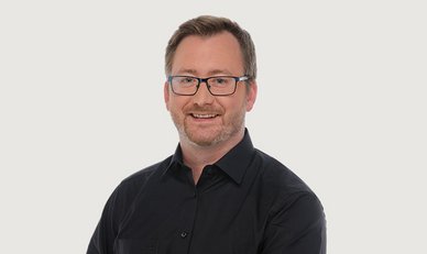 Der Mann mit einem gepflegten Bart und Brille strahlt Selbstbewusstsein aus, während er vor einem einfarbigen Hintergrund steht.