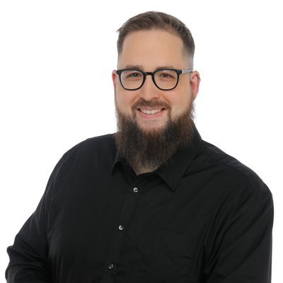 Ein lächelnder Mann mit Brille und Bart trägt ein schwarzes Hemd und posiert vor einem weißen Hintergrund.