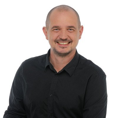 Mit einem selbstbewussten Lächeln präsentiert sich der Mann in einem schwarzen Oberteil vor einem hellen Hintergrund.